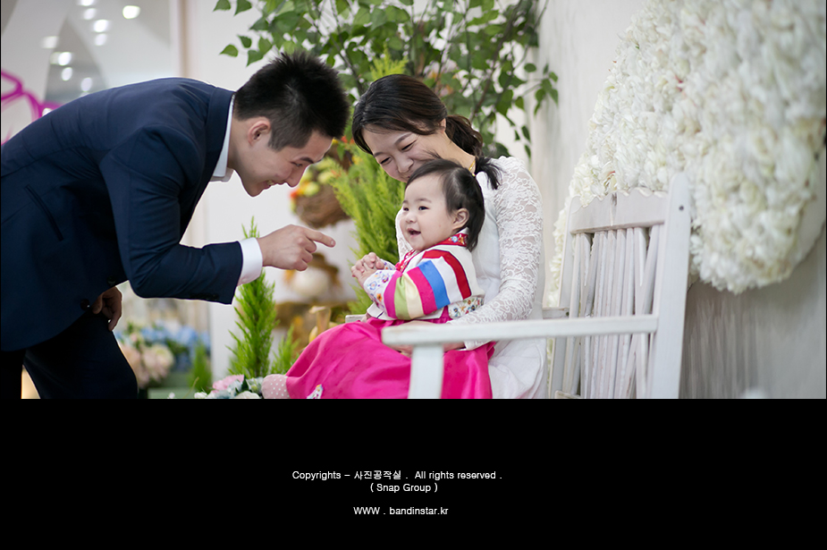 꽃 장식 앞 가족사진 - 아빠 손가락 향해 손 뻗는 아기