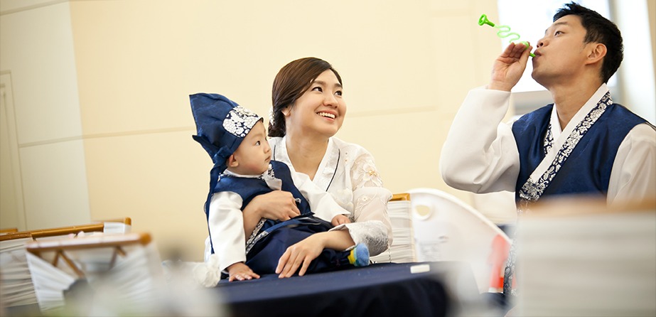 돌잔치홀 한복 차림 가족 3인 — 아빠가 장난감 피리를 불고 아이가 올려다보는 장면