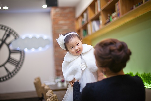 돌잔치홀 실내 — 엄마가 흰 드레스 아기를 들어 올려 마주보는 순간