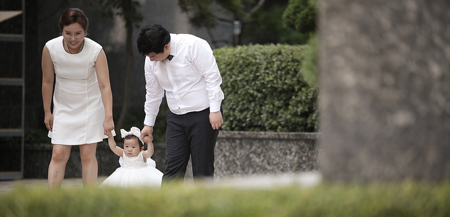 야외 정원 가족 3인 — 아빠가 하윤이 손 잡아 걸음마 돕고 엄마가 옆에서 바라보는 장면