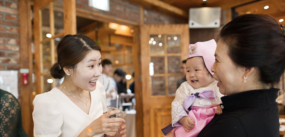 실내 하객 공간 — 엄마가 한복 차림 하윤이 안고 하객과 마주보며 웃는 장면