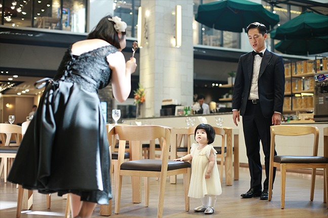 돌잔치홀 내부 — 아이가 의자 사이에 혼자 서 있고 부모가 양쪽에서 바라보는 장면